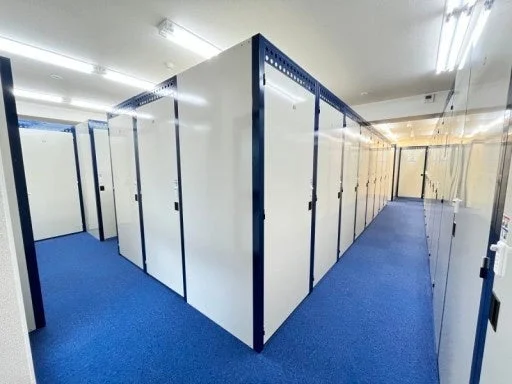 青いカーペットが敷かれた明るい室内空間に、白い扉と濃い青色のフレームを持つ多数の貸し倉庫(トランクルーム)が整然と並ぶ廊下の様子です。