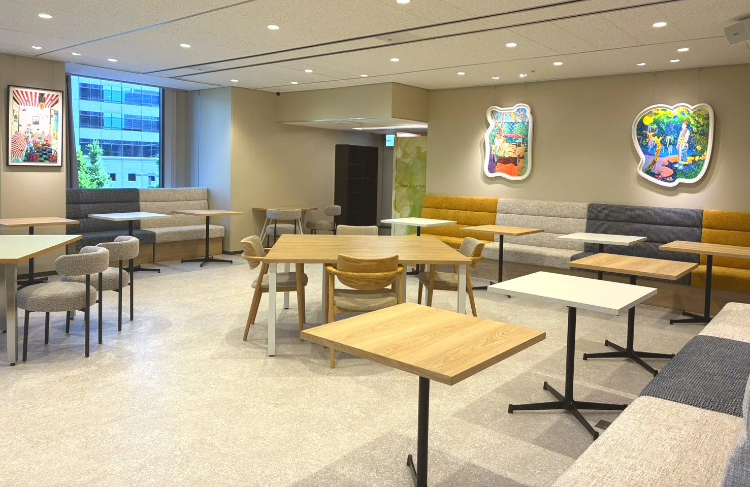 明るくモダンな雰囲気のオフィスの休憩室またはカフェテリア。様々な形状のテーブルと椅子、カラフルなソファ席が配置され、壁にはアート作品が飾られた快適な空間です。